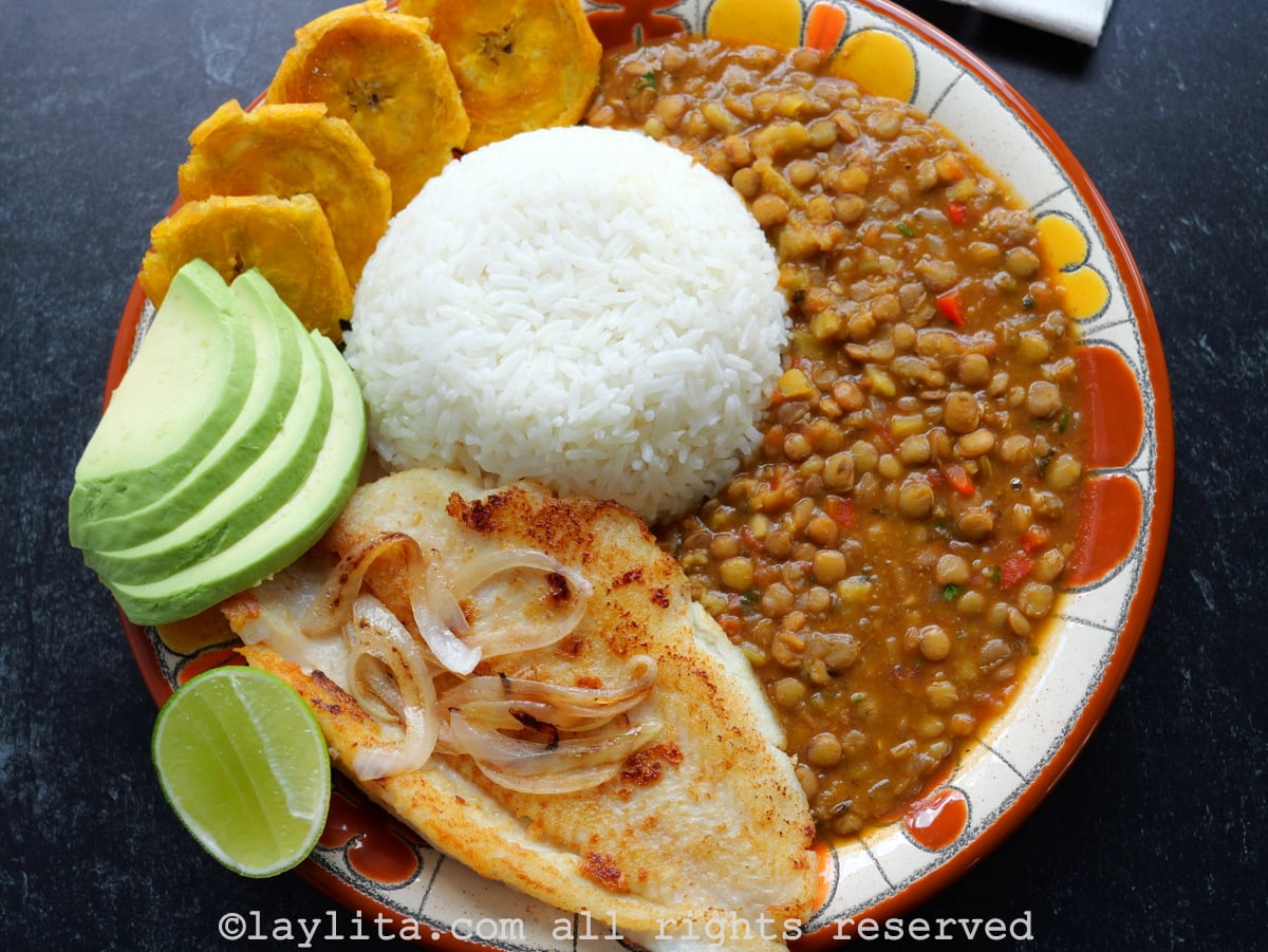 A brown orange floral plate filled with lentils, rice, plantains, fish, avocado and a half lime