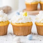 Three coconut cupcakes topped with coconut frosting, coconut flakes and easter egg candies on a kitchen counter with egg candies and coconut flakes on the side