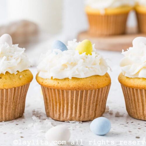 Three coconut cupcakes topped with coconut frosting, coconut flakes and easter egg candies on a kitchen counter with egg candies and coconut flakes on the side