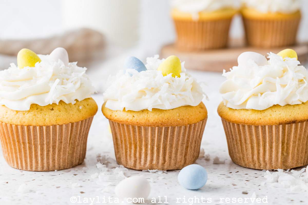 Three coconut cupcakes topped with coconut frosting, coconut flakes and easter egg candies on a kitchen counter with egg candies and coconut flakes on the side