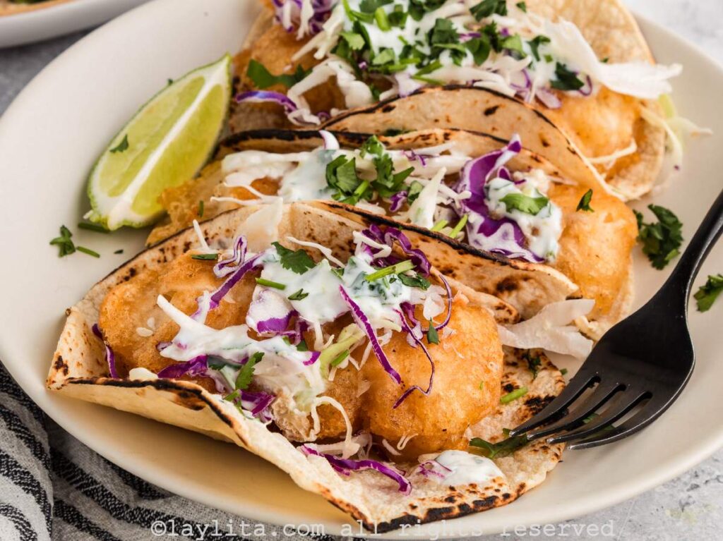 Close up of three Baja Fish tacos on a white plate, the tacos are topped with coleslaw and cilantro lime crema