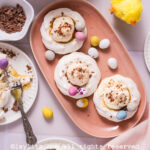 A pink platter with three meringue nests filled with cream and decorated with mini chocolate eggs, next to a white plate with a meringue nest and a fork breaking it apart