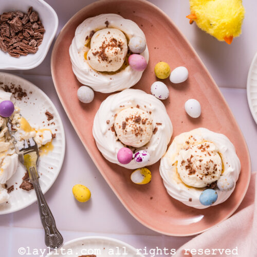 A pink platter with three meringue nests filled with cream and decorated with mini chocolate eggs, next to a white plate with a meringue nest and a fork breaking it apart