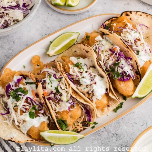 Overhead view of a platter of five Baja Fish tacos topped with slaw and Mexican crema, with lime wedges on the side