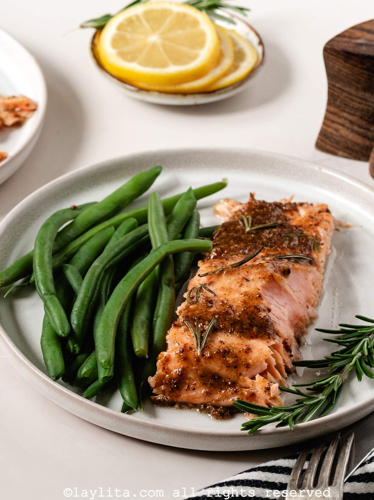 Sideview of a glazed roasted salmon fillet with green beans on a white plate, with lemon slices in the backgroun
