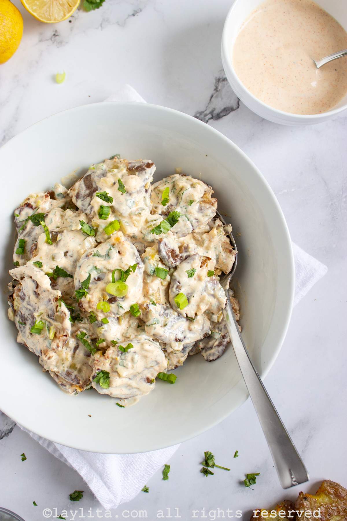 Overhead shot of smashed potato salad on a plate next to two smashed potatoes on the side and a bowl of creamy sauce in the background