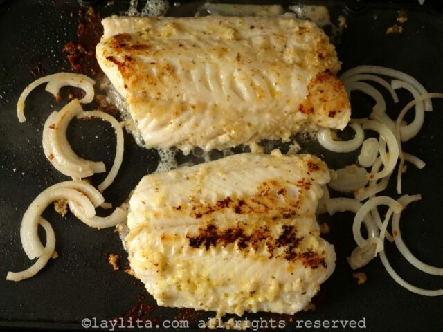 Two fish fillets on a hot gridle with onions cooking next to the fish