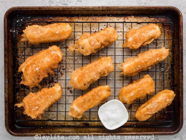 Fried battered cod fish strips on a wire rack with a small bowl of salt