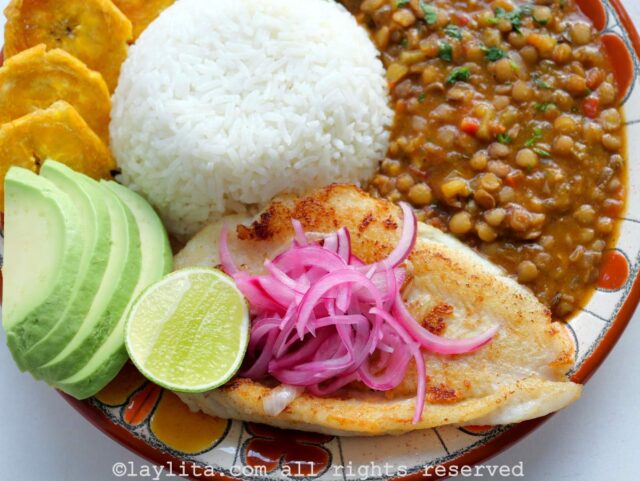Close up of pescado a la plancha style fish on a plate with rice and lentils, with red onions on top of the fish.