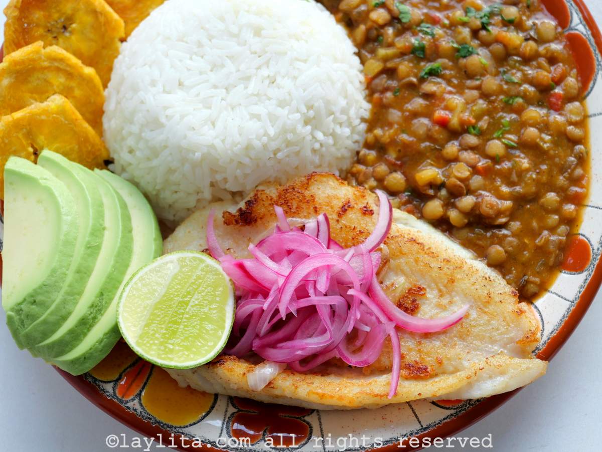Close up of pescado a la plancha style fish on a plate with rice and lentils, with red onions on top of the fish.