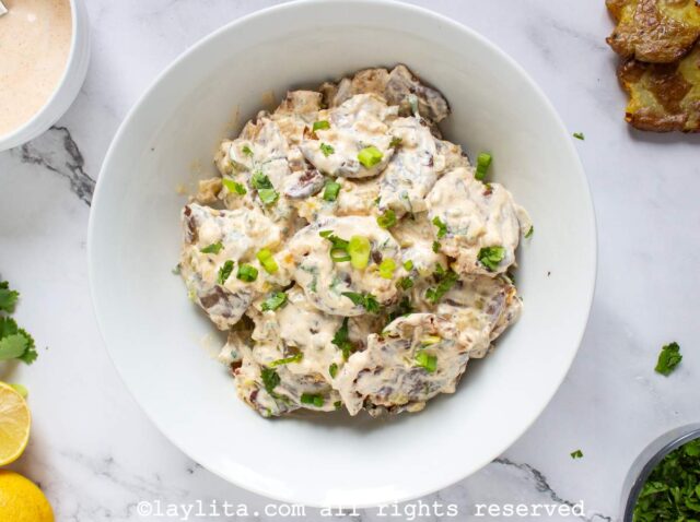 Overhead shot of crispy smashed potato salad in a bowl next to a small bowl of dressing, a half cut lemon and a couple of smashed potatoes