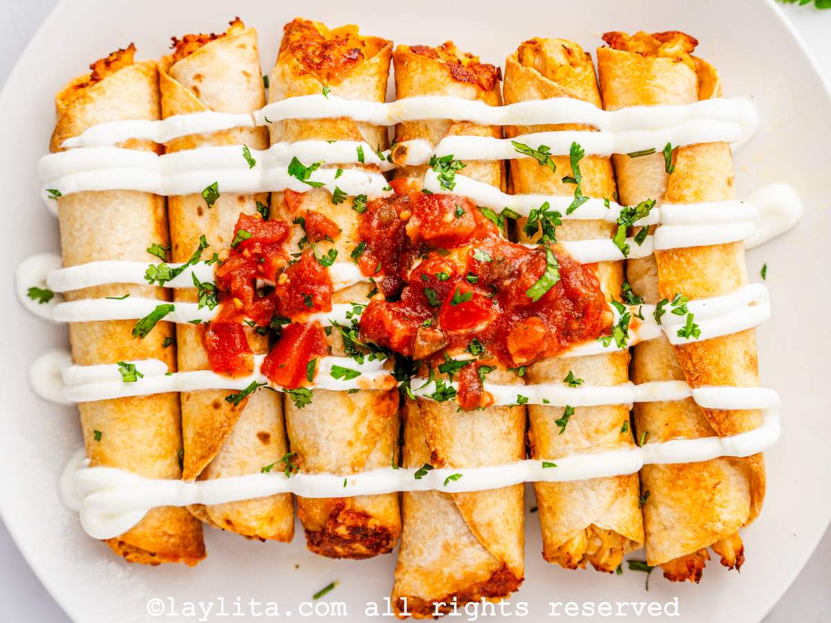 Air fryer chicken taquitos on a white plate with sour cream and salsa on top.