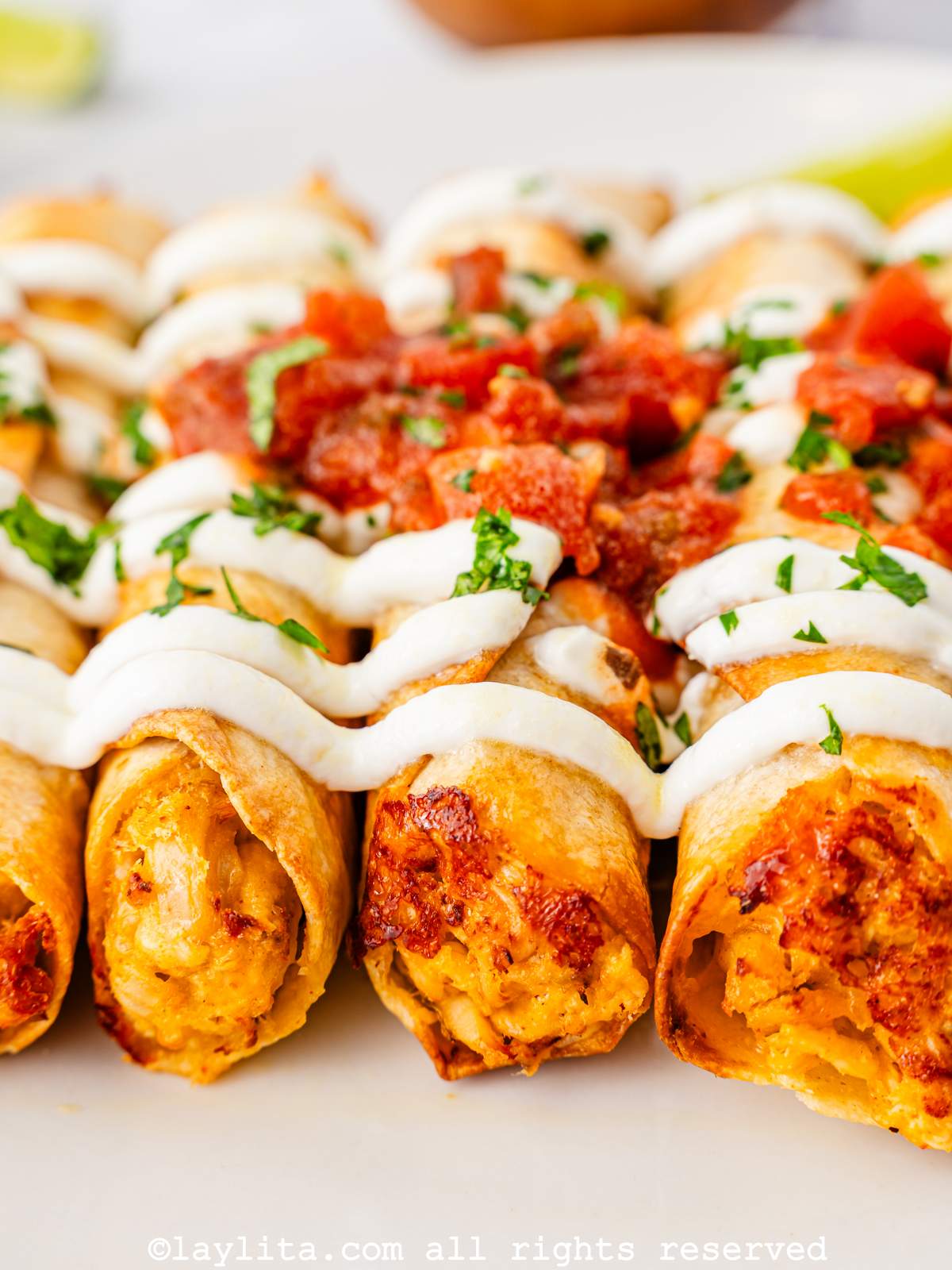 Close up of air fryer chicken taquitos lined up on a plate garnished with Mexican crema and salsa.