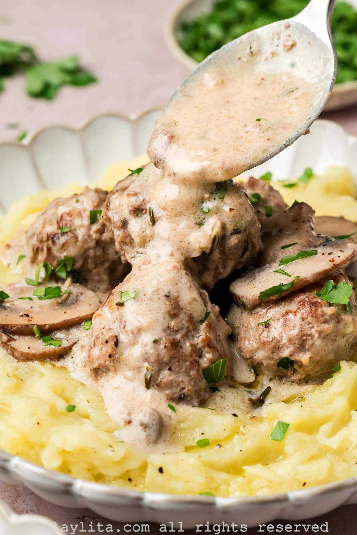 Mashed potatoes and beef stroganoff meatballs in a bowl with a spoon drizzling sauce on top.
