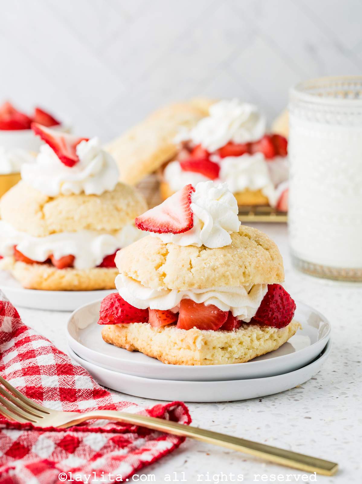 A strawberry shortcake on a plate with several more in the background, next to a red and white kitchen napkin and a fork