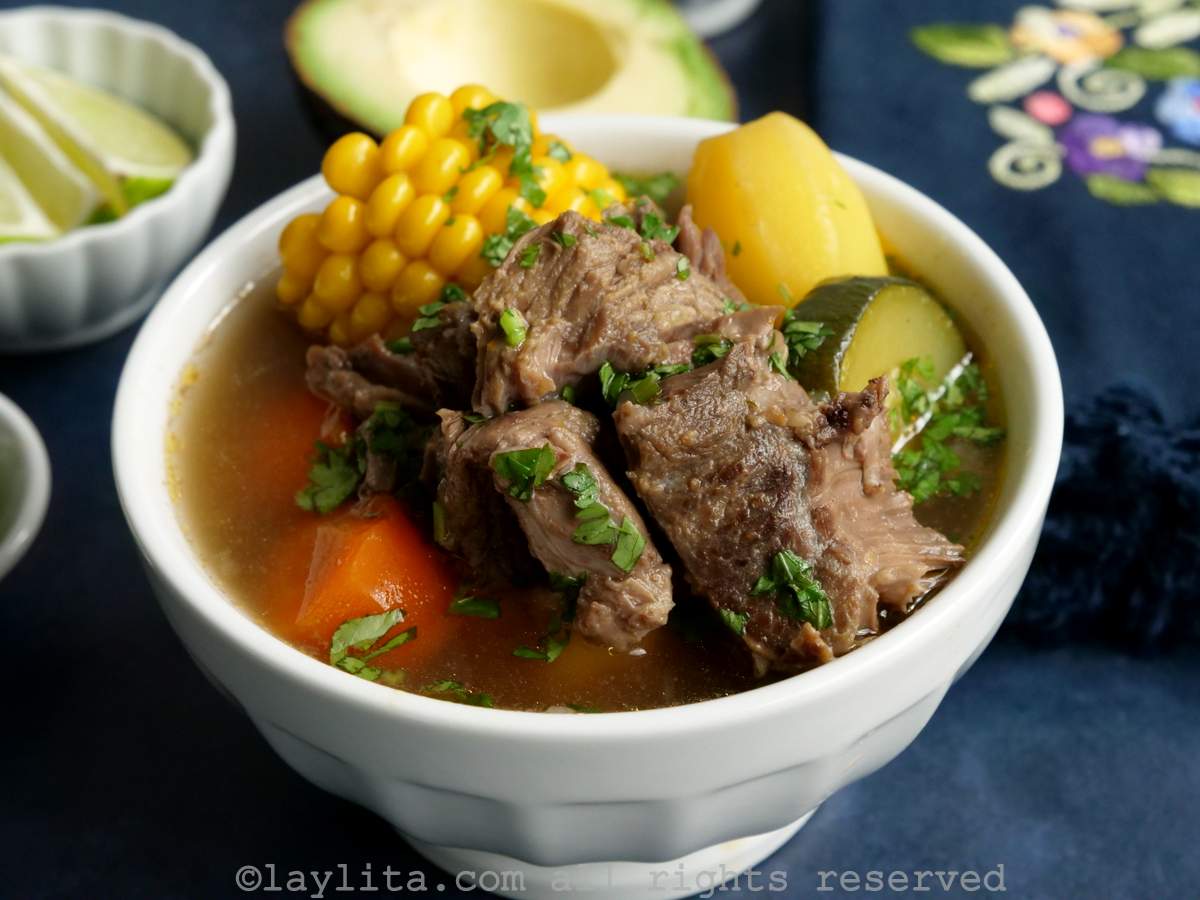 A white soup bowl full of Mexican beef soup with chunks of beef and vegetables, with avocado and lime slices in the background