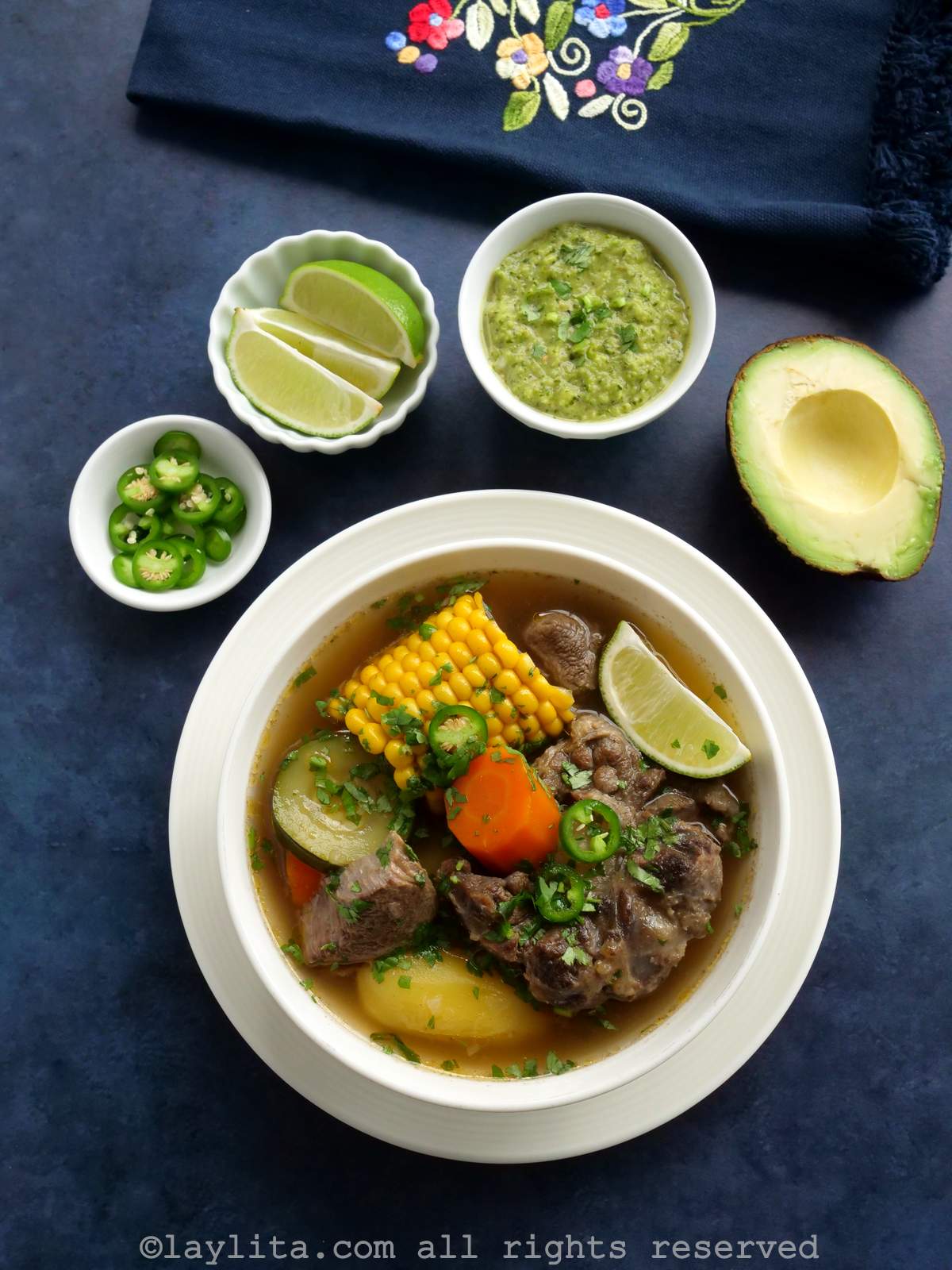 Overhead shot of caldo de res in a white bowl on a blue background with garnishes on the side.