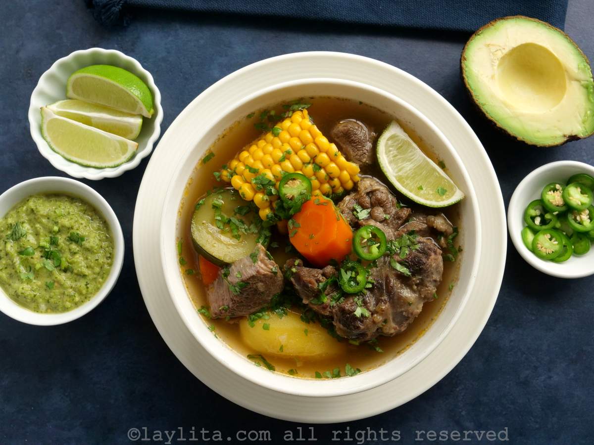 Overhead shot of Mexican caldo de res beef soup in a white bowl on a blue background with garnishes on both sides