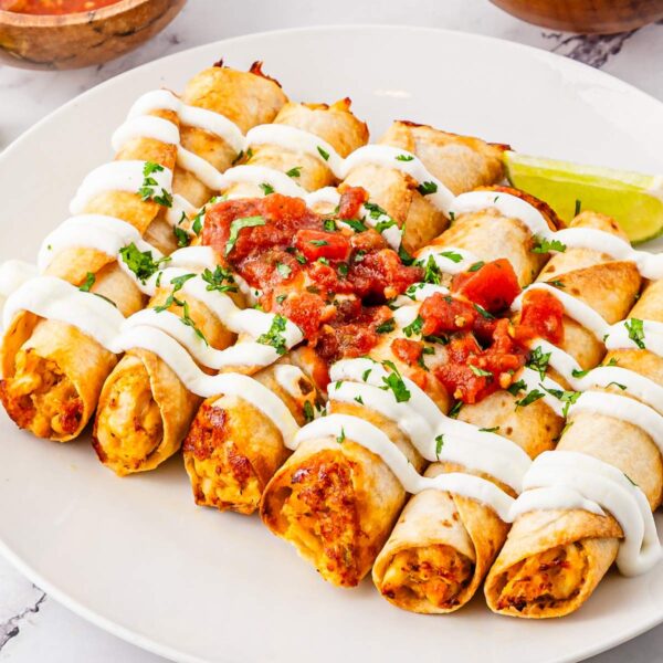 A white plated with several crispy taquitos or flautas garnished with Mexican crema and red salsa