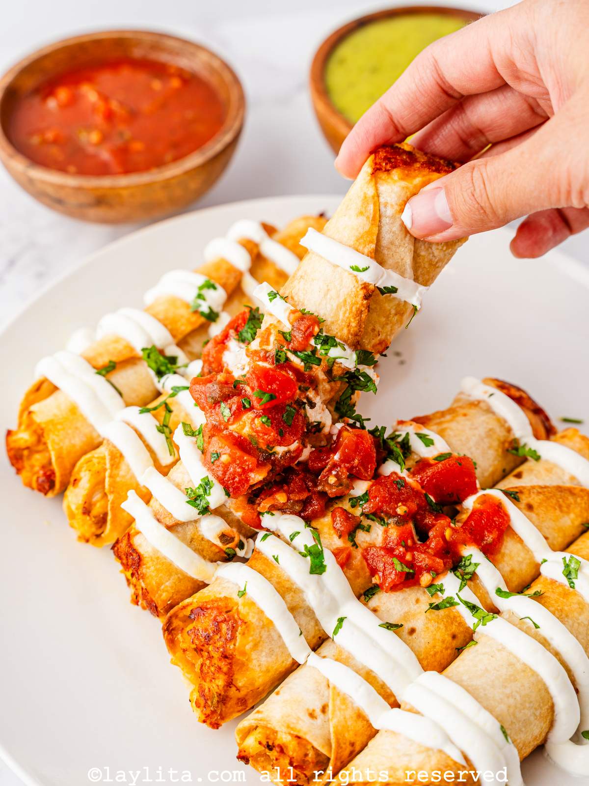 Air fryer chicken taquitos on a plate with a hand taking one, with some dips in the background