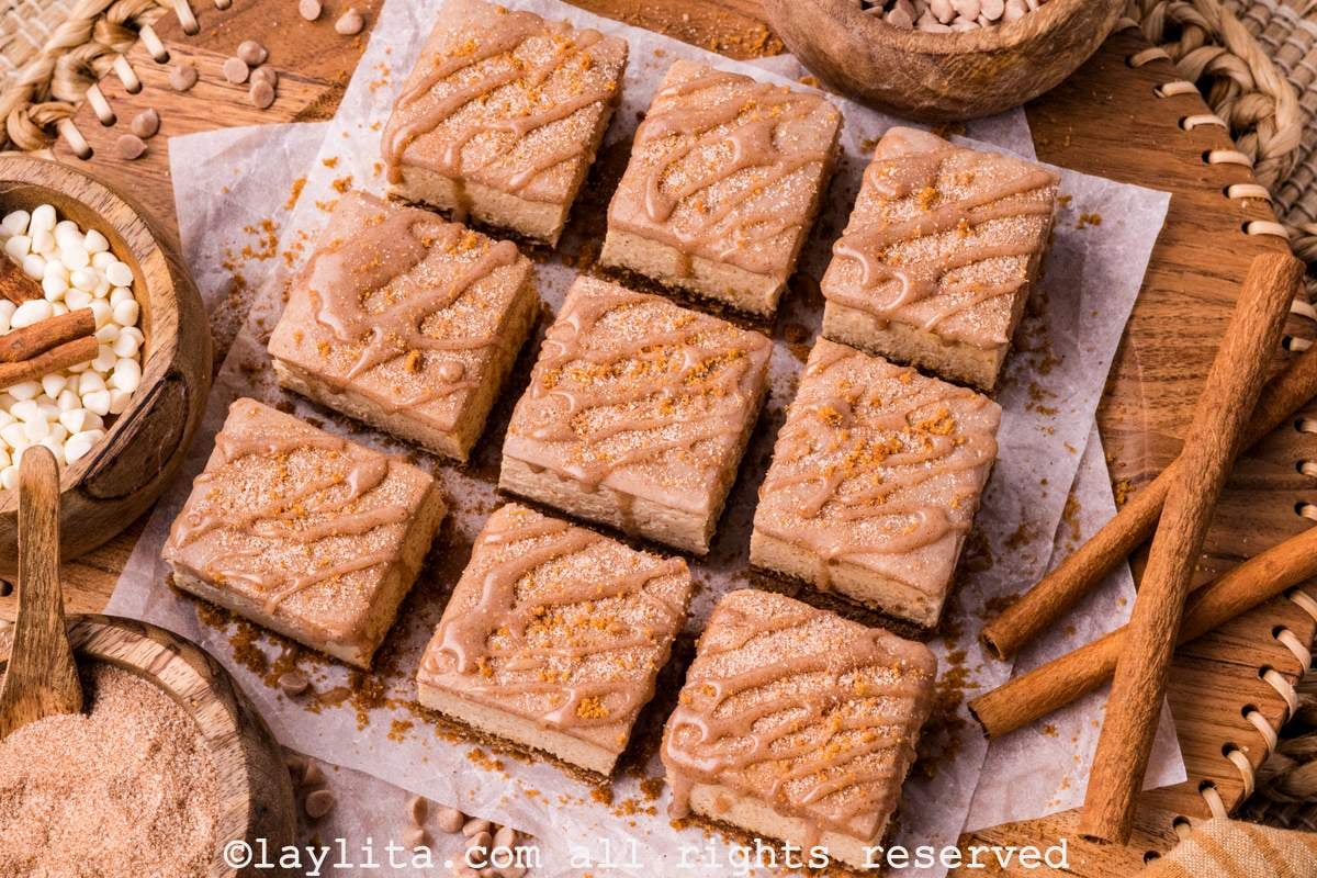 Ovrehead view of nine churro cheesecake bars on parchment paper with cinnamon sticks and cinnamon chips on the side