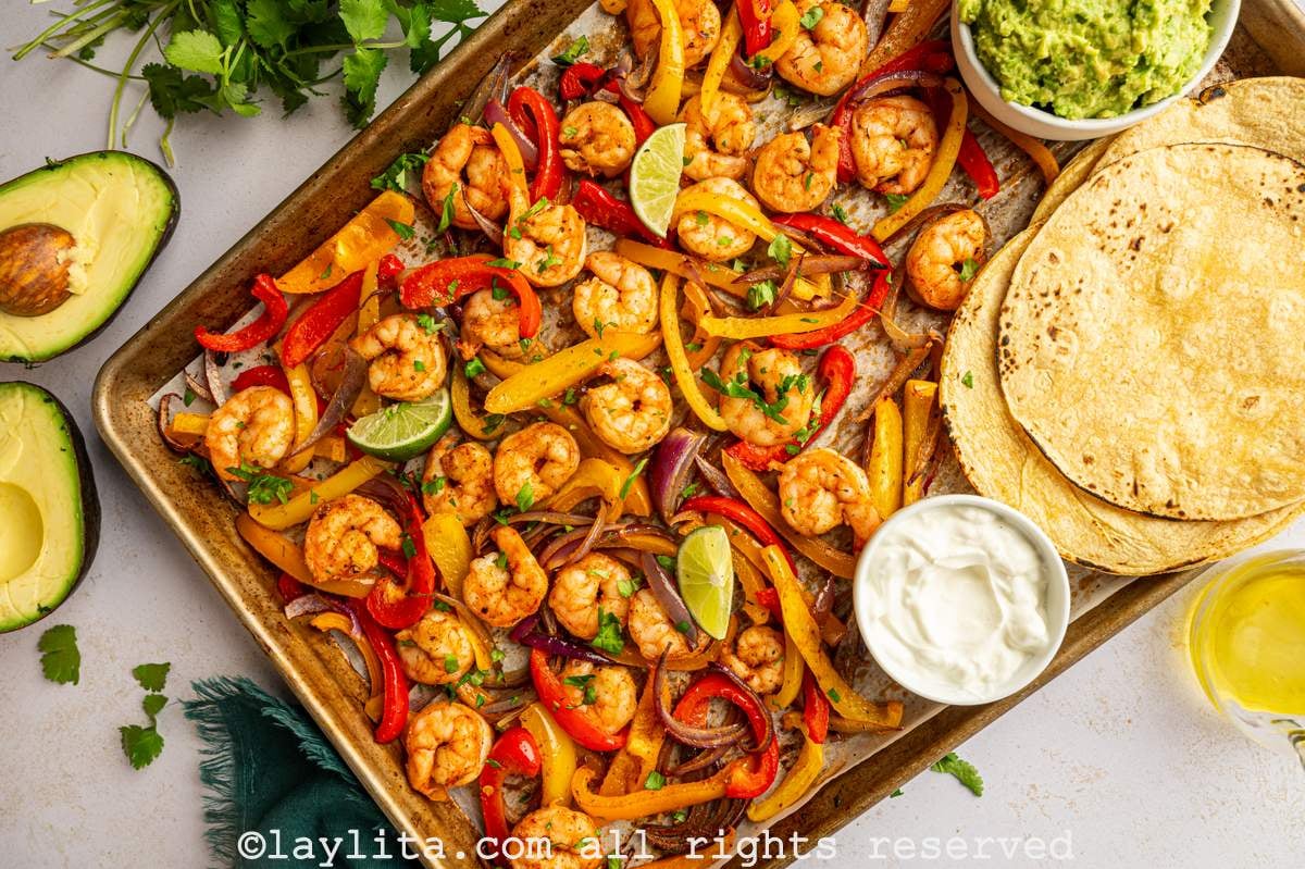 View of a baking sheet pan with Shrimp Fajitas next to a stack of corn tortillas, a bowl of guacamole and sour cream. On the left side there are some cilantro sprigs and an avocado cut in half.