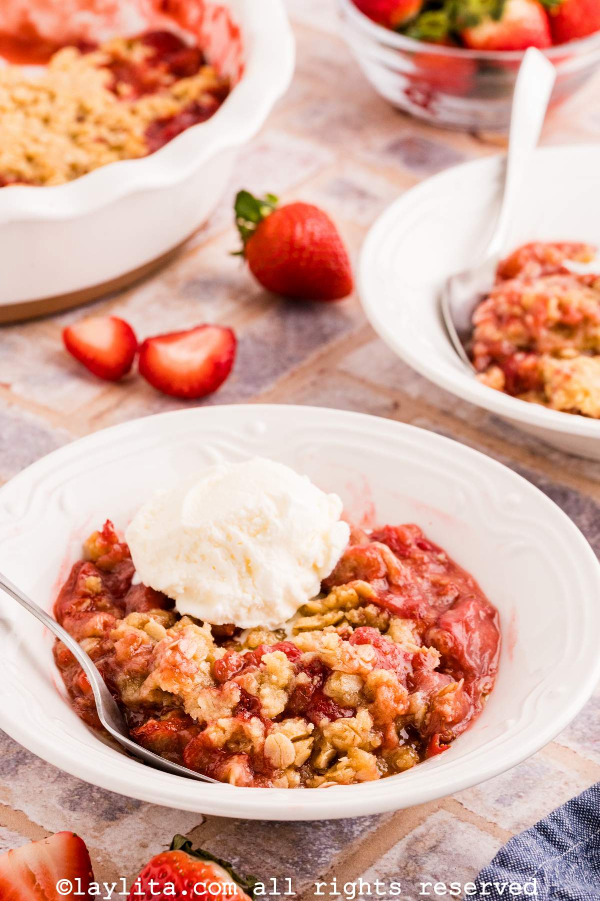 A portion of strawberry crisp in a white bowl with a scoop of vanilla ice cream, with more strawberry crisp in the background