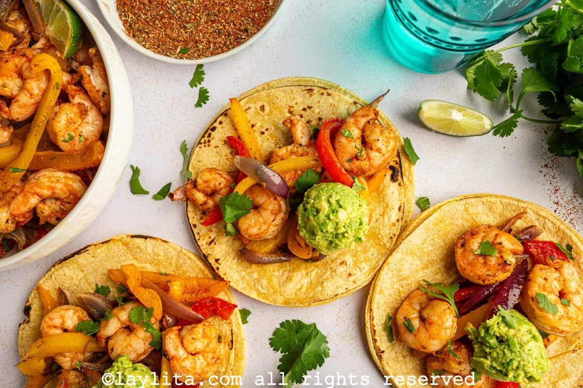 Three shrimp fajita tacos topped with mashed avocado, with a bowl of shrimp fajitas, extra avocado, lime wedges and cilantro on the side