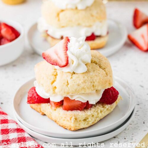 Two plates of strawberry shortcakes lined up, both garnished with strawberries and cream., with additional strawberries on the side