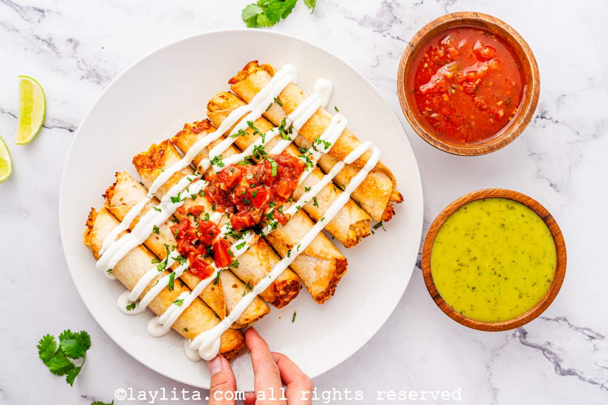 Overhead shot of air fryer chicken flautas or taquitos on a plate with a hand taking one, next to some dips