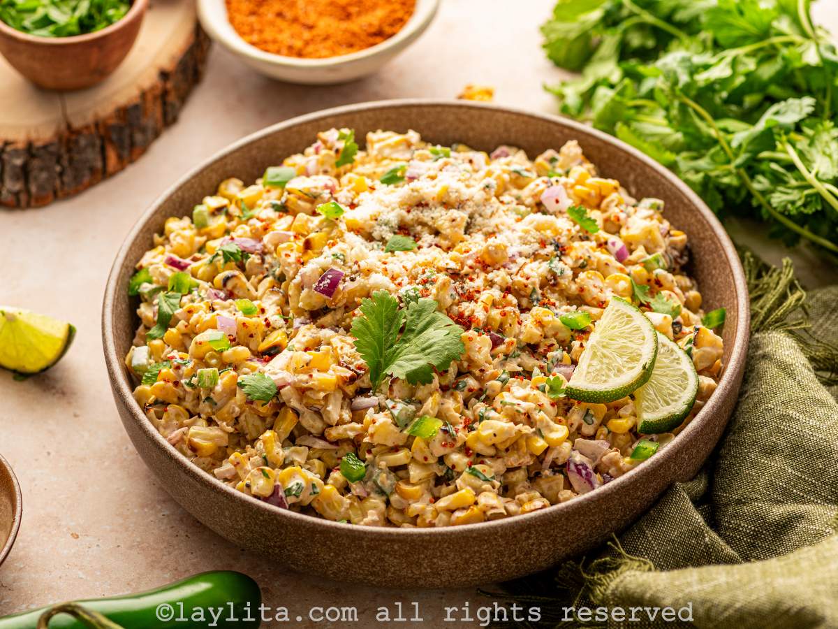A large brown bowl filled with Mexican corn salad, with a couple of lime slices on top. In the background there is a small bowl of chili powder and some cilantro sprigs