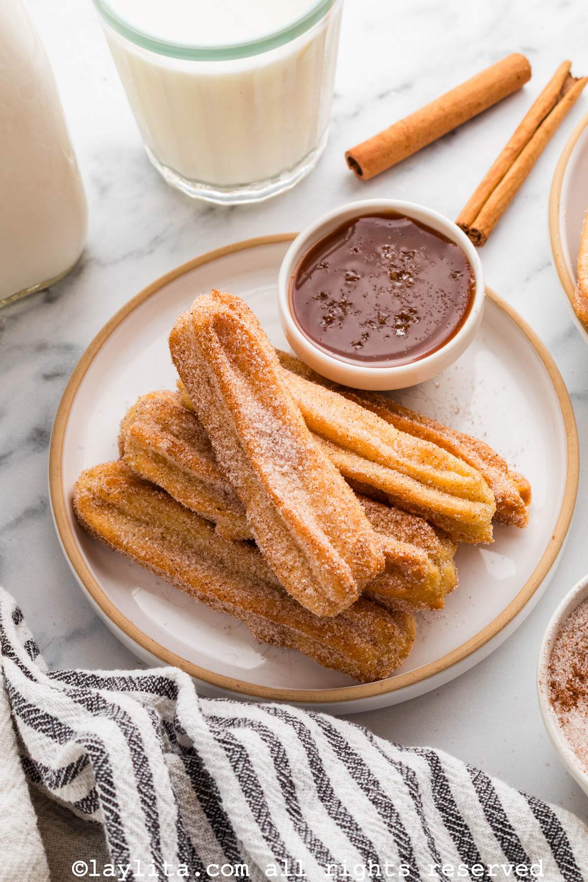 A plate with several churros with a bowl of caramel sauce next to them and a glass of milk in the background