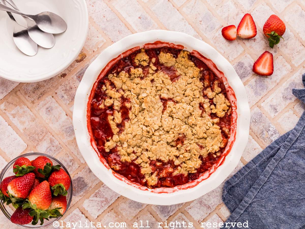 A tile countertop with a pie dish full of strawberry dessert in the center, on the sides there are fresh strawberries, a blue kitchen towel and a white plate with three spoons on it