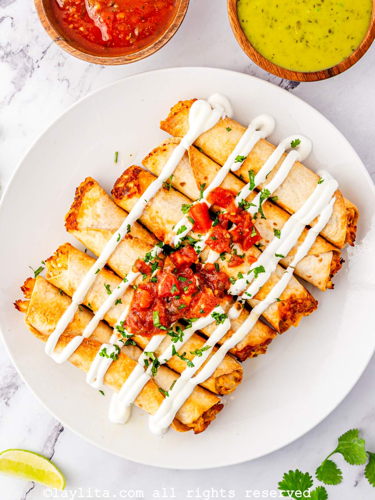 A white plated with several crispy taquitos or flautas garnished with Mexican crema and red salsa, with two bowls of salsa on the side