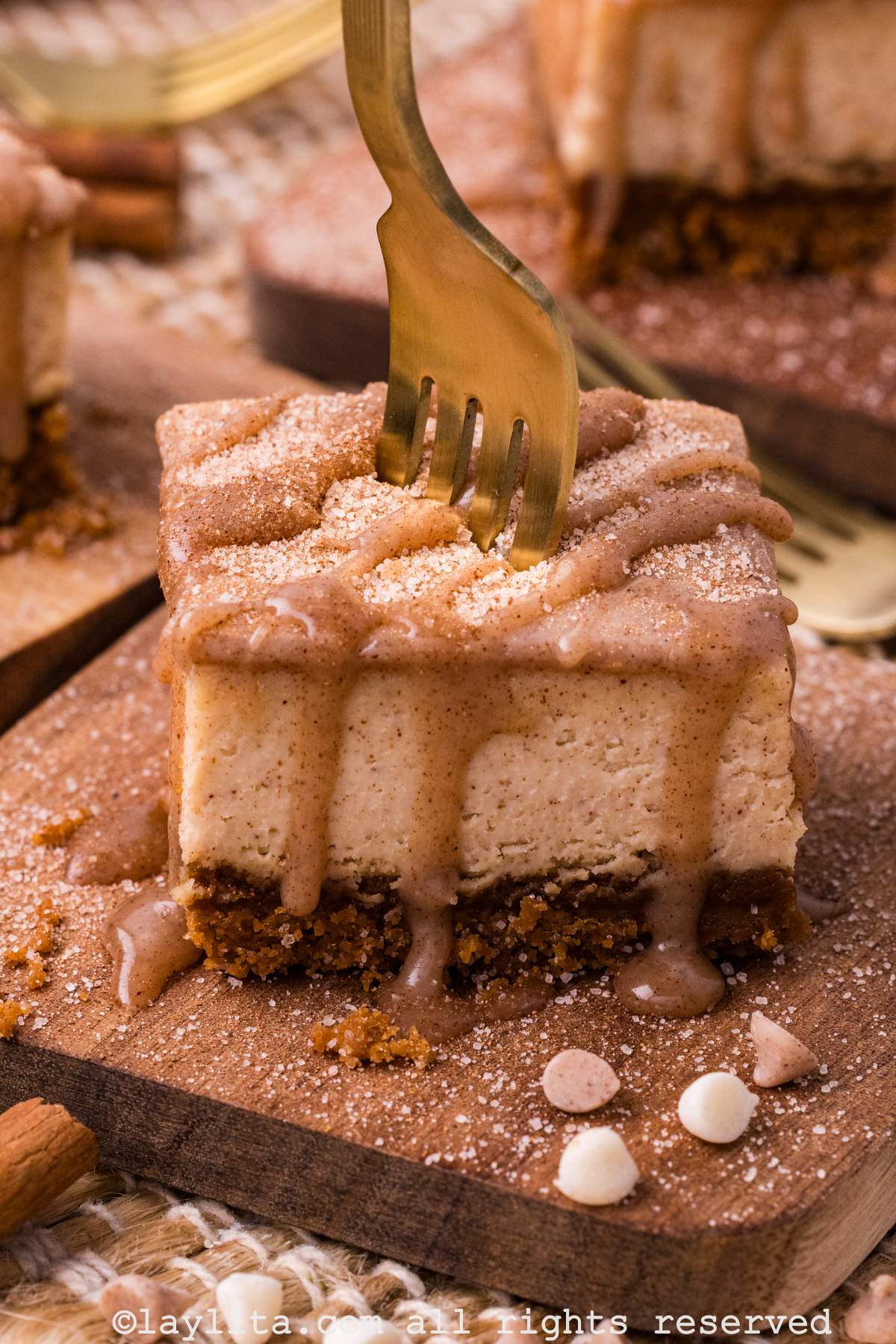 A square piece of cheesecake topped with vanilla ganache on a wooden board, with a fork in the middle of the cheesecake