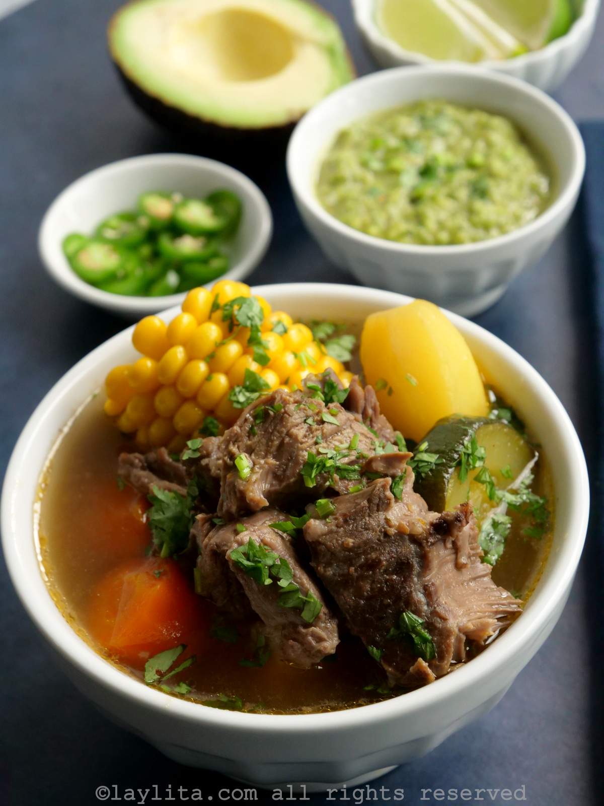 Side view of a white bowl of Mexican caldo de res with garnishes in the background