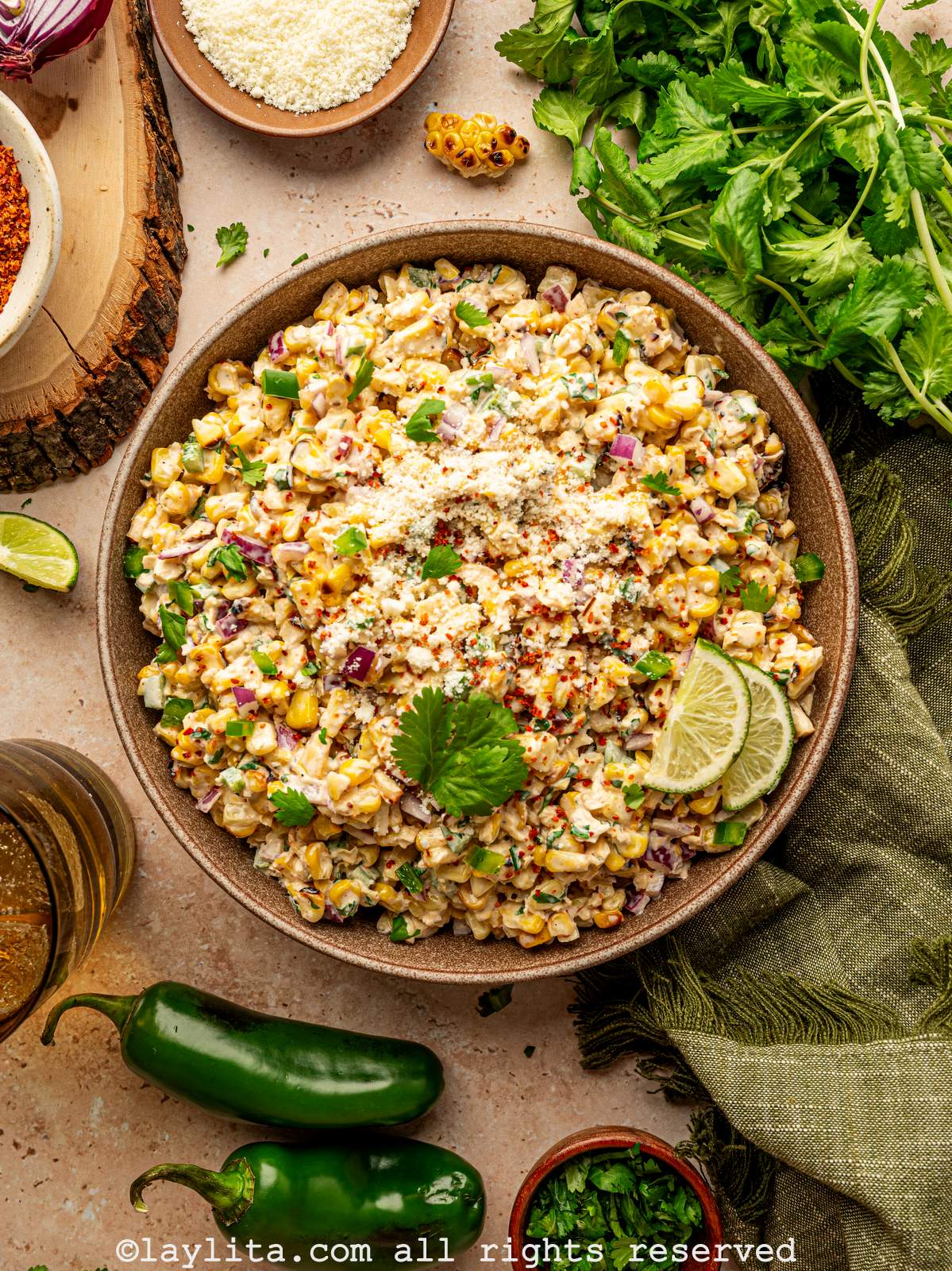 Overhead view of a bowl of corn salad topped with cilantro, crumbled cheese and lime slices, with several garnishes and green kitchen towel on the side