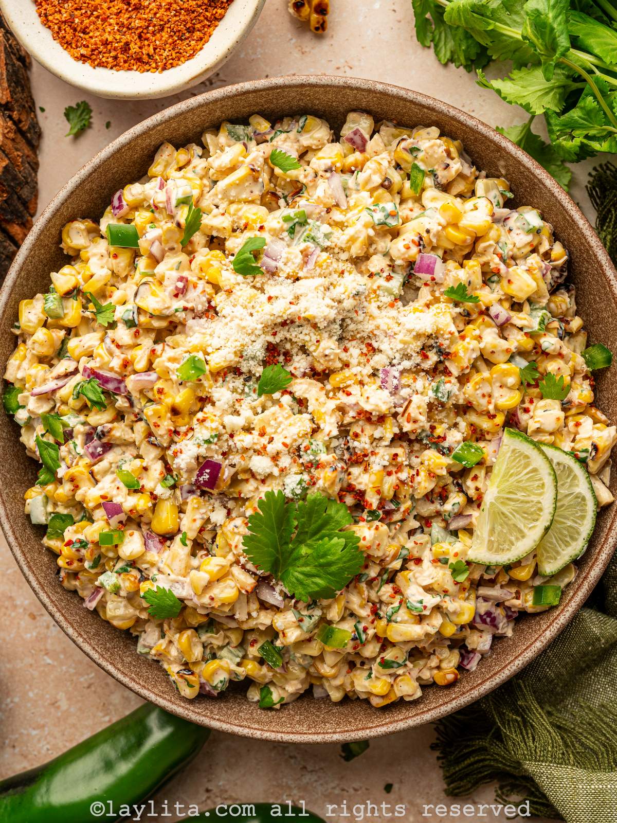 Closeup shot of a bowl of Mexican street corn salad with cilantro leaves and lime slices on top, in the background there is a bowl of chili powder