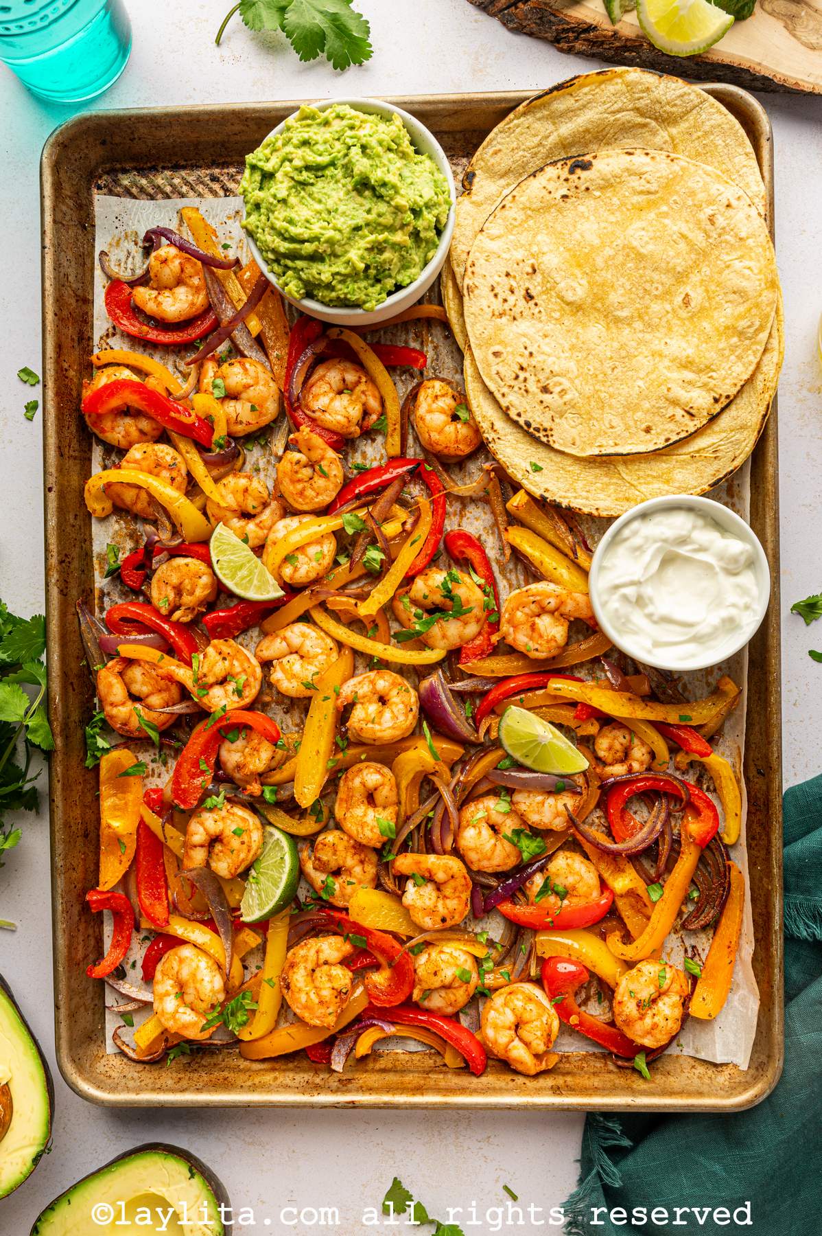 A baking sheet pan filled with roasted shrimp and sliced veggies, with corn tortillas, mashed avocado and sour cream on the side of the pan