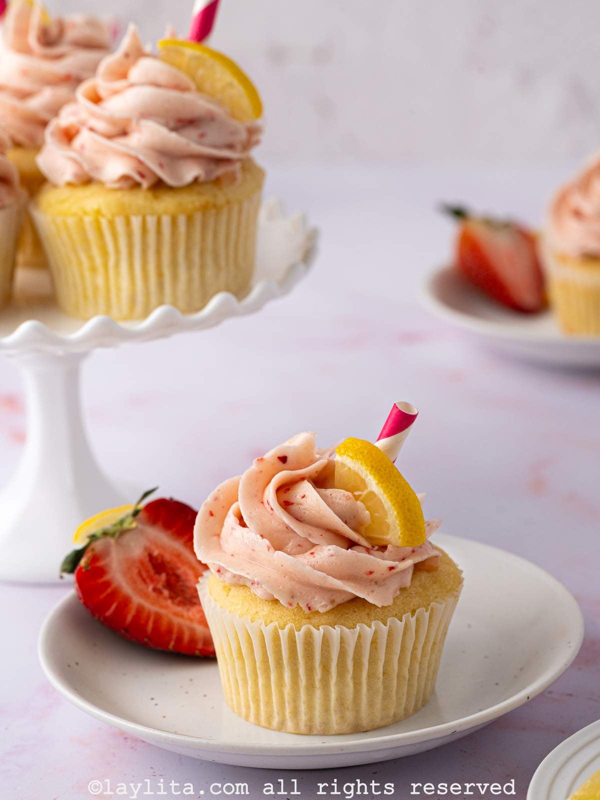 A strawberry lemonade cupcake decorated with a small decorate striped straw on a white plate with more cupcakes on a stand in the background