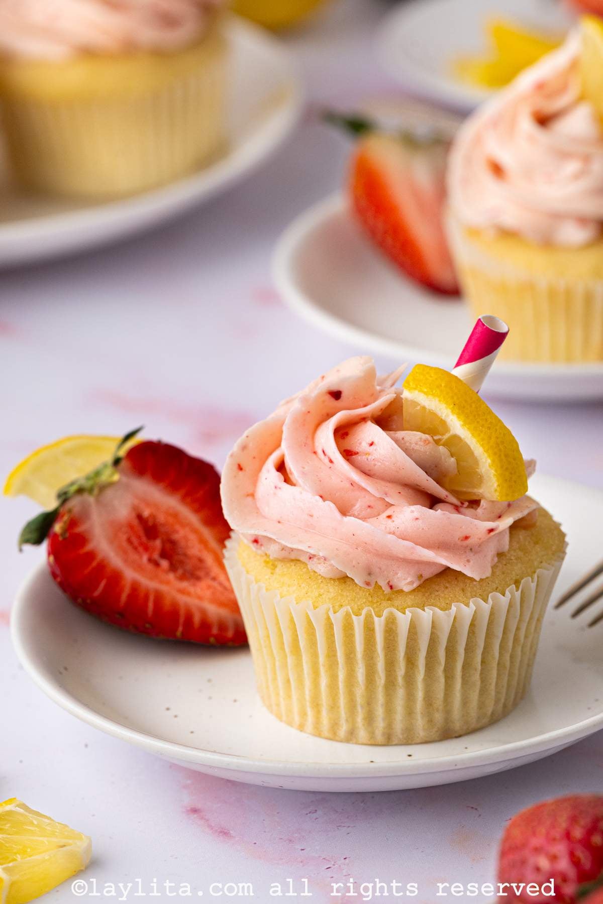 A lemon cupcake topped with strawberry buttercream frosting on a white plate, with more cupcakes in the background