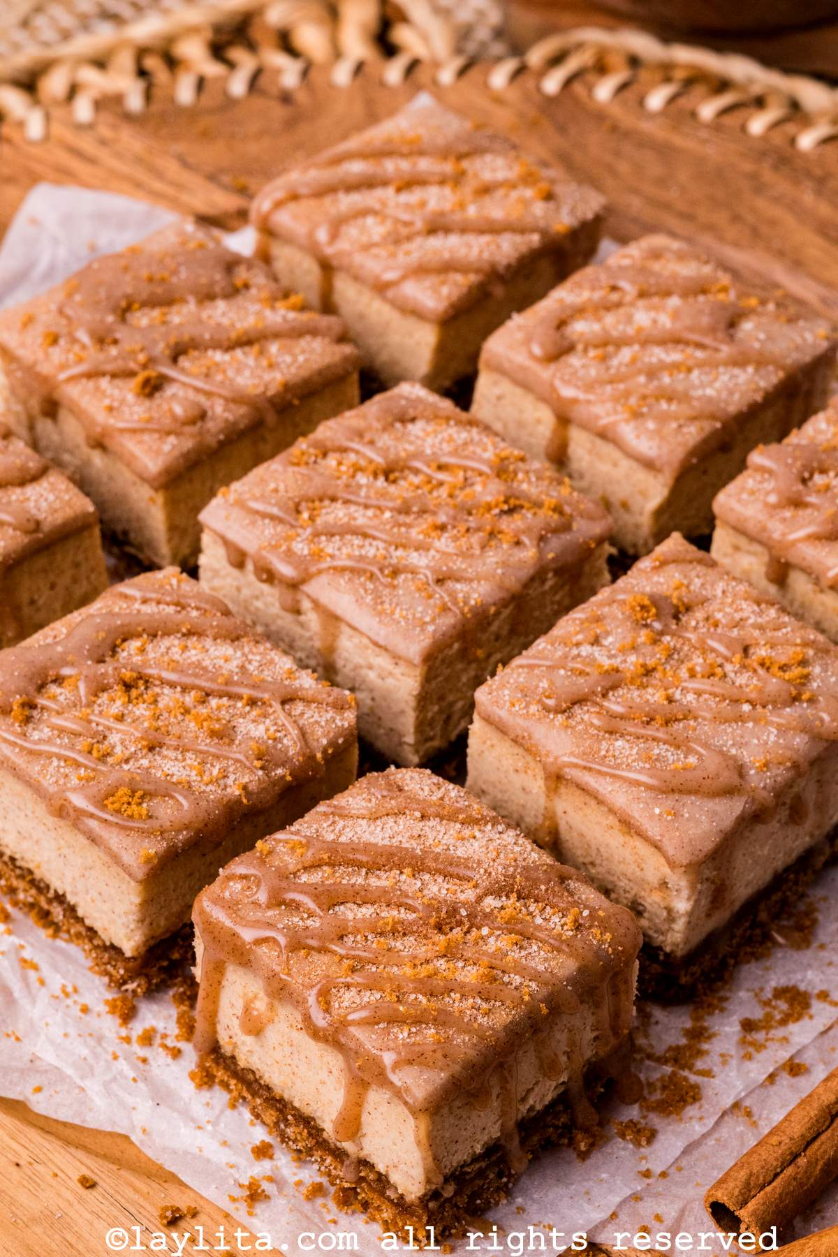 A churro cheesecake cut into nine bars, topped with cinnamon ganache, on a piece of parchment paper with cinnamon sticks and chips on the side