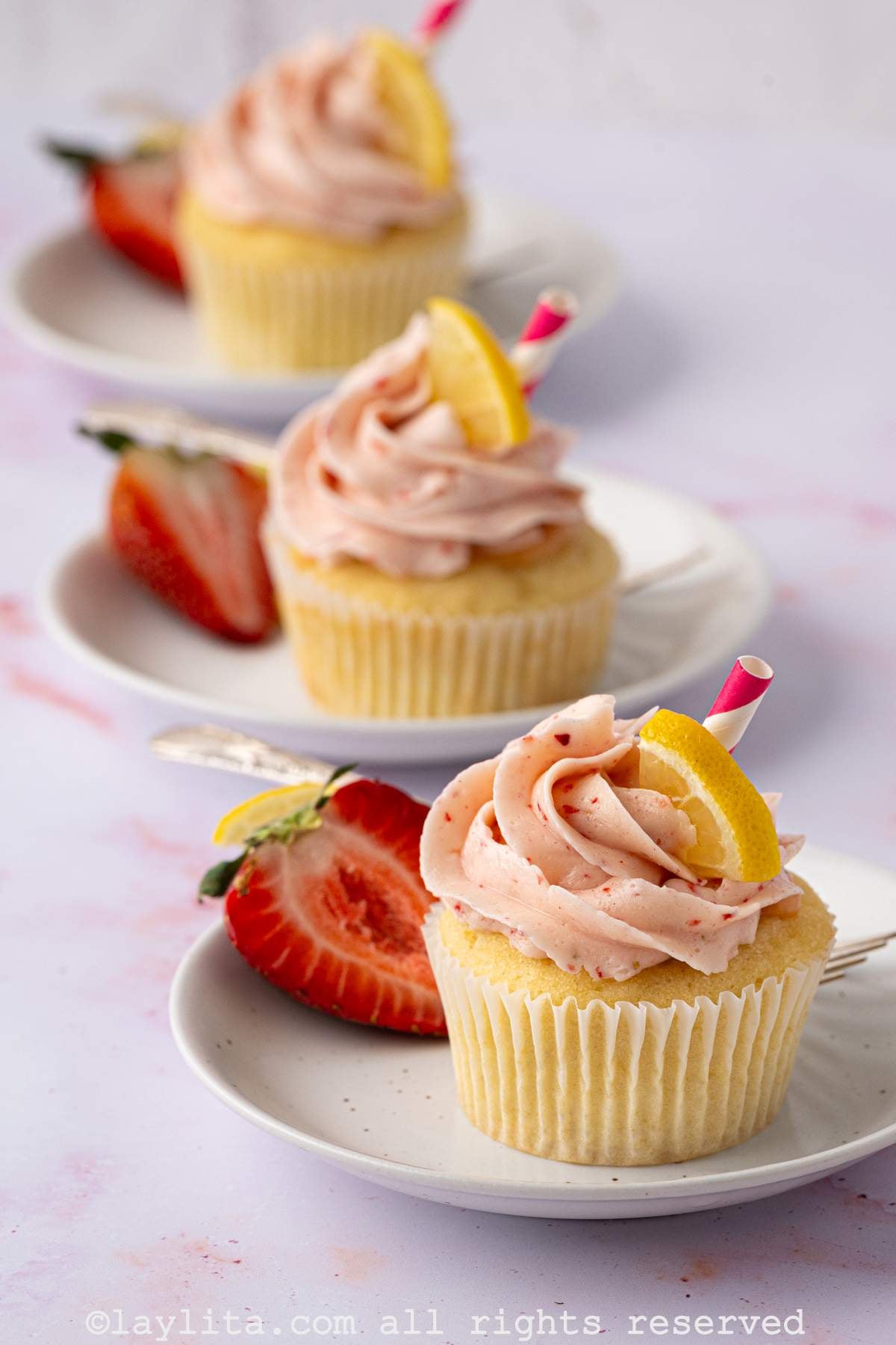 Three strawberry lemonade cupcakes lined up, each one is on its own white plate