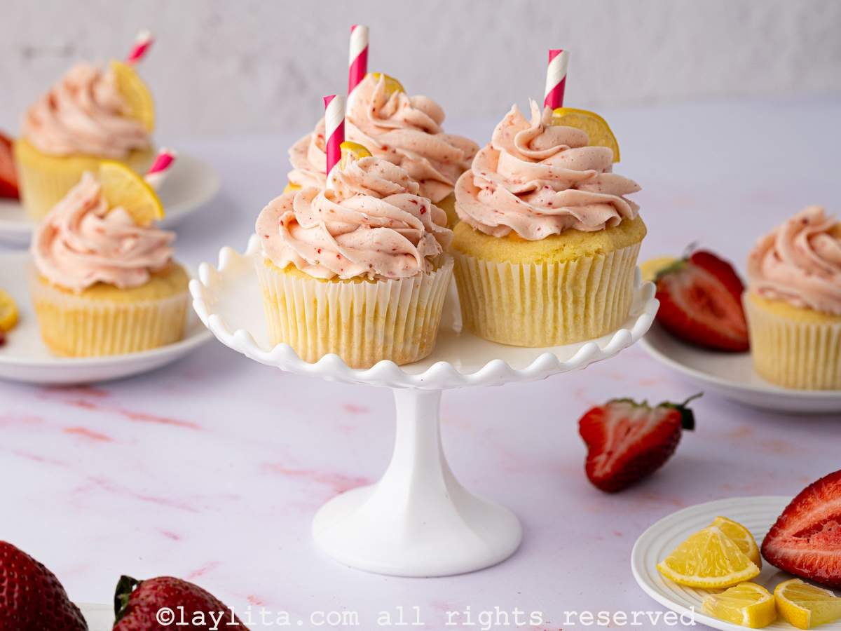 Sideview of three strawberry lemon cupcakes on a small white cake stand, with more cupcakes and strawberries in the background