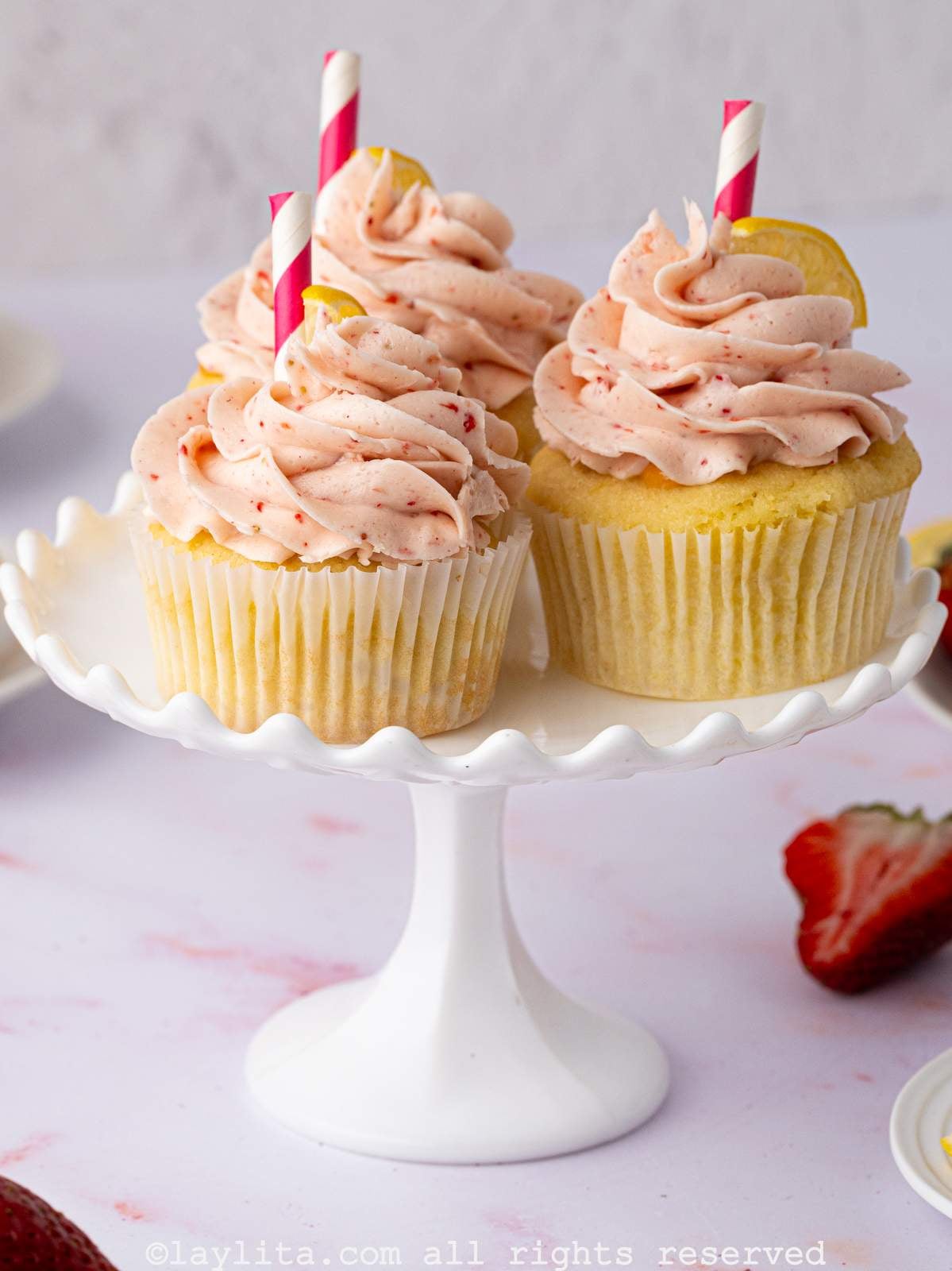 Three strawberry lemon cupcakes on a small white cake stand, with strawberries in the background