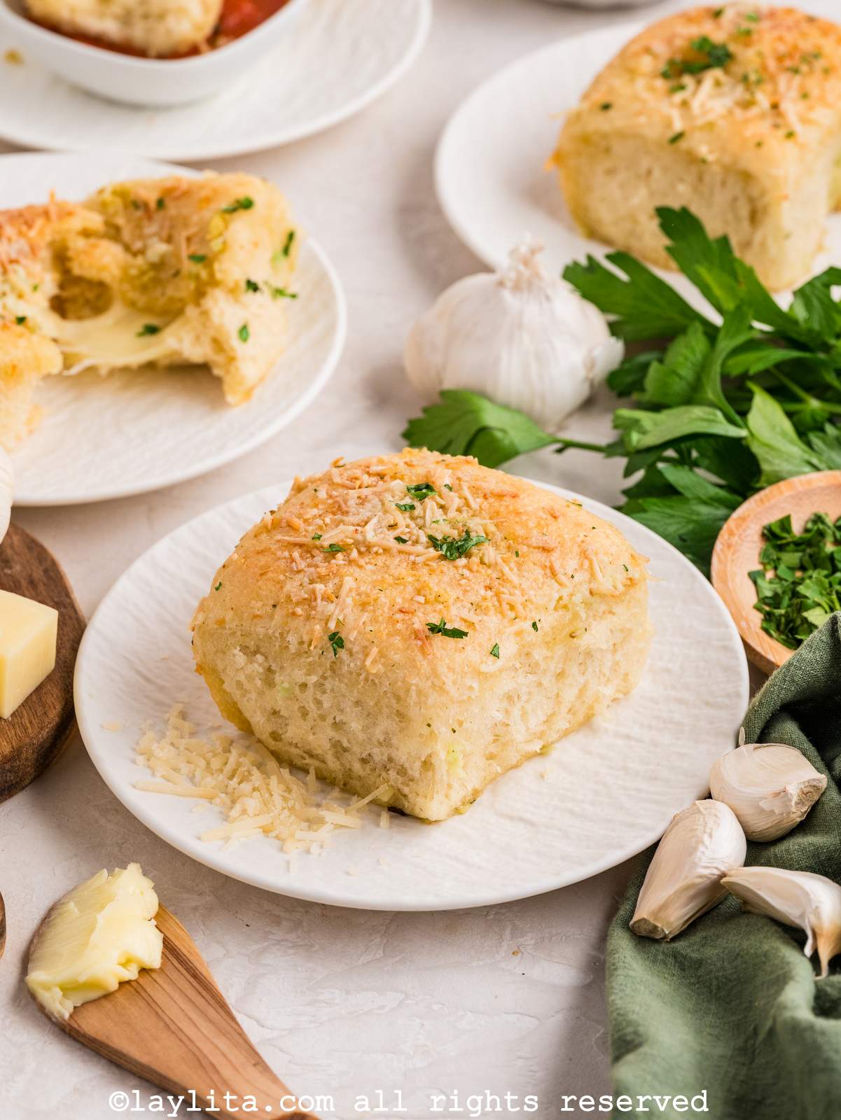 A cheesy bread roll topped with parmesan and chopped parsley on a white plate with more bread rolls in the background