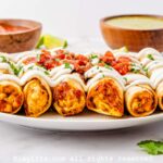 Side view of air fryer chicken taquitos lined up on a plate garnished with sour cream and salsa.
