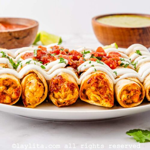 Side view of air fryer chicken taquitos lined up on a plate garnished with sour cream and salsa.