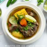 Overhead shot of caldo de res in a white bowl with lime, avocado and jalapeño salsa on the side