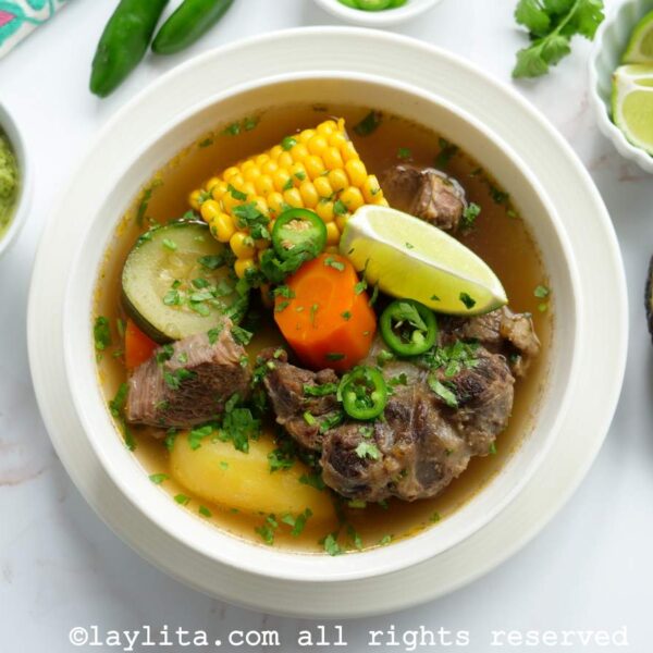 Overhead shot of caldo de res in a white bowl with lime, avocado and jalapeño salsa on the side